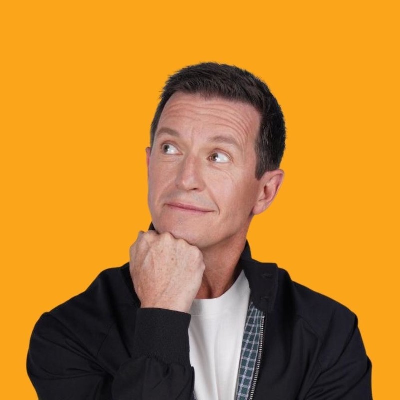 Mid shot of Rove McManus wearing a black jacket with blue checked lining and a white t-shirt. He has his chin resting on his fist and is looking up to his right. Yellow background.