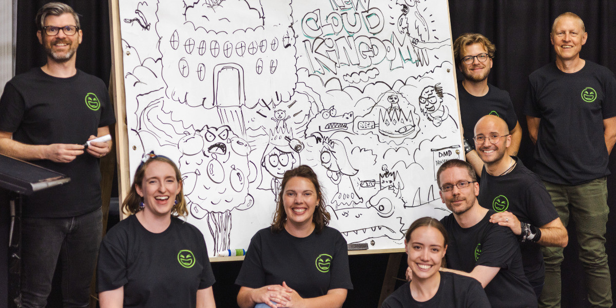 Group shot of smiling cast and crew in matching black t-shirts in front of a whiteboard filled with silly cartoons.
