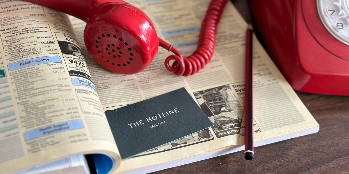 The Hotline - The receiver and cord for a red rotary phone lies on top of an open Yellow Pages. A pencil and a plain green business card also sits on the page. The card reads: The Hotline. Call now.