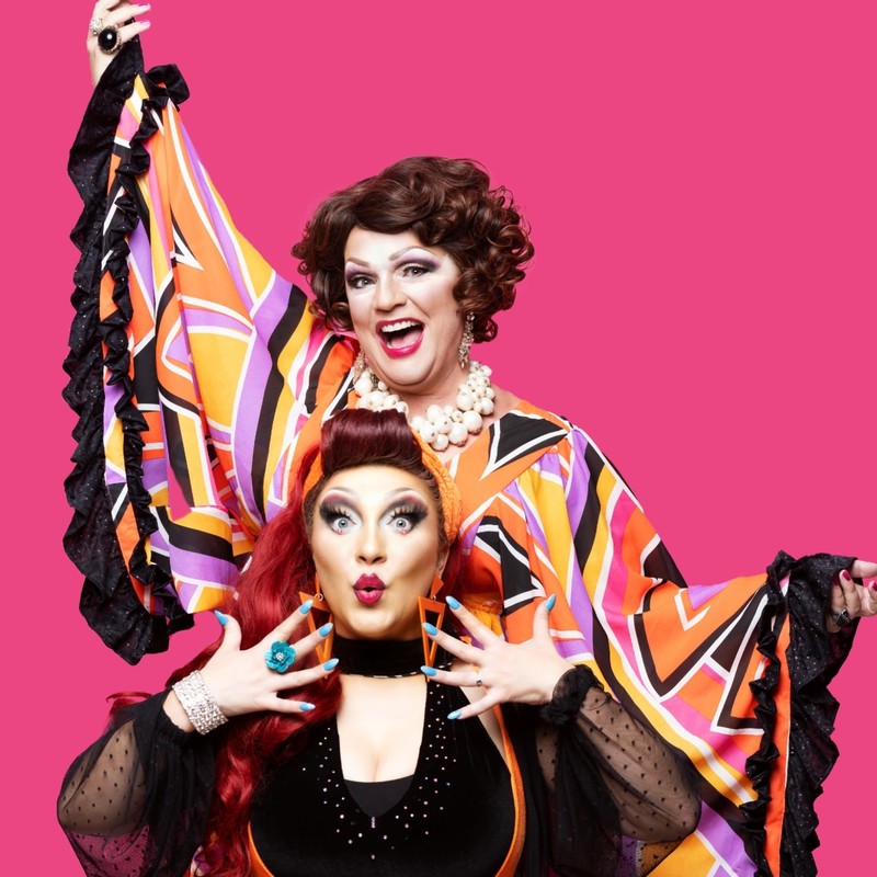 Two brightly coloured drag queens are smiling and posing to camera with pink background.