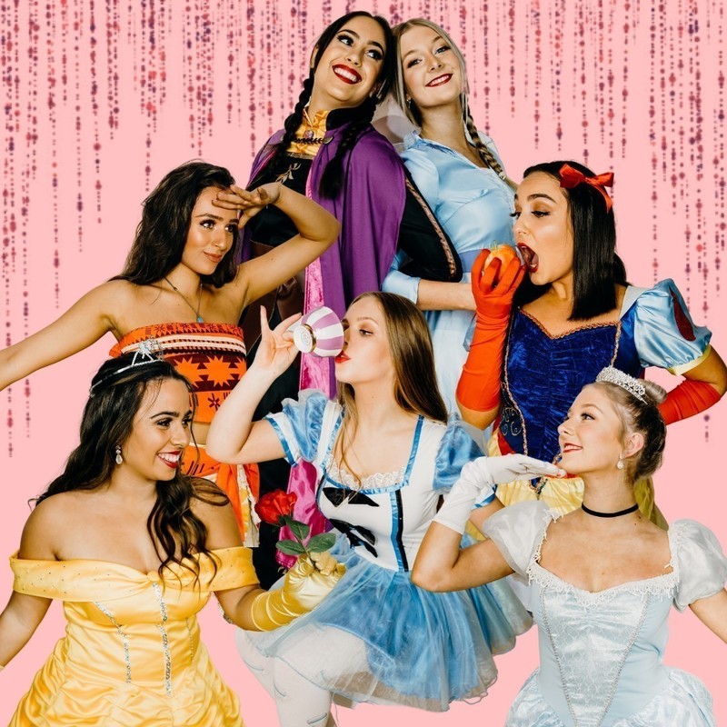 Seven women dressed as disney princesses are standing together. At the top are two princesses, one in a purple cape and brunette braids the other in a blue dress and blond braids. In the middle are three princesses, from left to right - one has brown hair and an patterned orange bandeau top, the middle is blond in a blue and white dress with a tea cup, the third has brown is in a blue dress, orange gloves and is about to eat an apple. At the bottom level are two princesses, one with brown hair and a yellow off shoulder gown, the other blonde in a pale blue dress, white gloves and wearing a tiara.