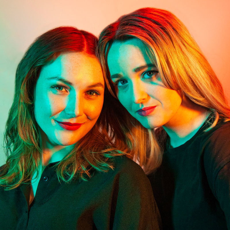Different Now - Image Description: Two women dressed in black shirts and jeans stand close together. They look into the camera and smile. They stand against a blank wall and are lit from the sides with bright green and orange lighting.