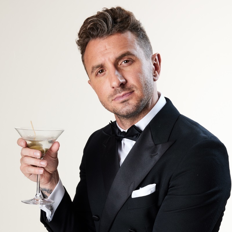 Tommy looking at the camera, wearing a tuxedo and bowtie and holding a martini glass