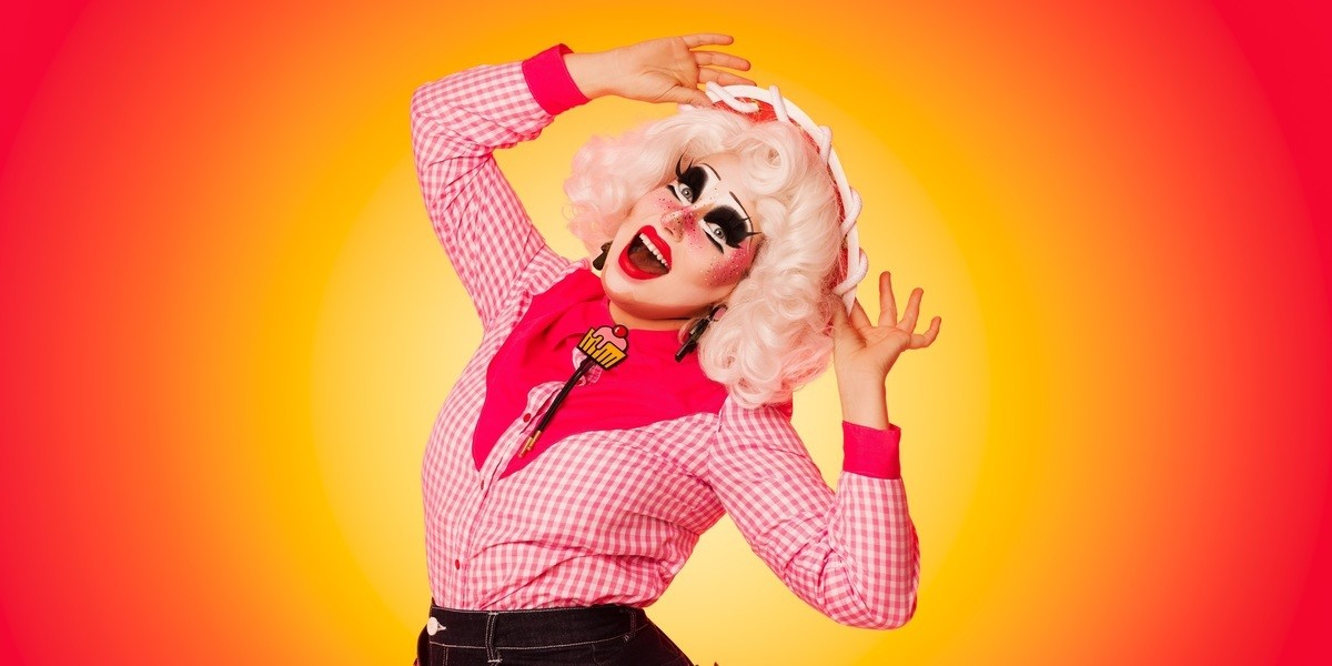 The drag queen Clara Cupcakes is smiling at the camera and tilting her hat to the viewer while standing in front of a sunset gradient background. She is dressed as a cowgirl in a red hat with white trim, a pink checked shirt with a pink cupcake bolo tie and denim shorts. Her hair is white and she has a full face of drag make-up.