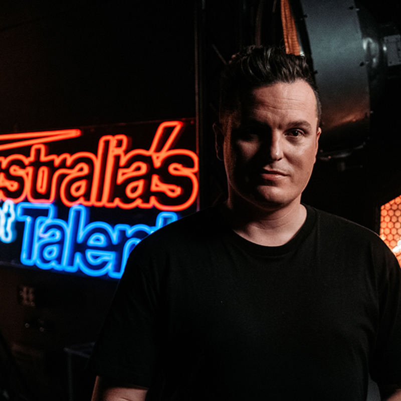 Matt Tarrant wearing a black t-shirt and facing the camera with bright lights and a neon sign of Australias Got Talent in the background