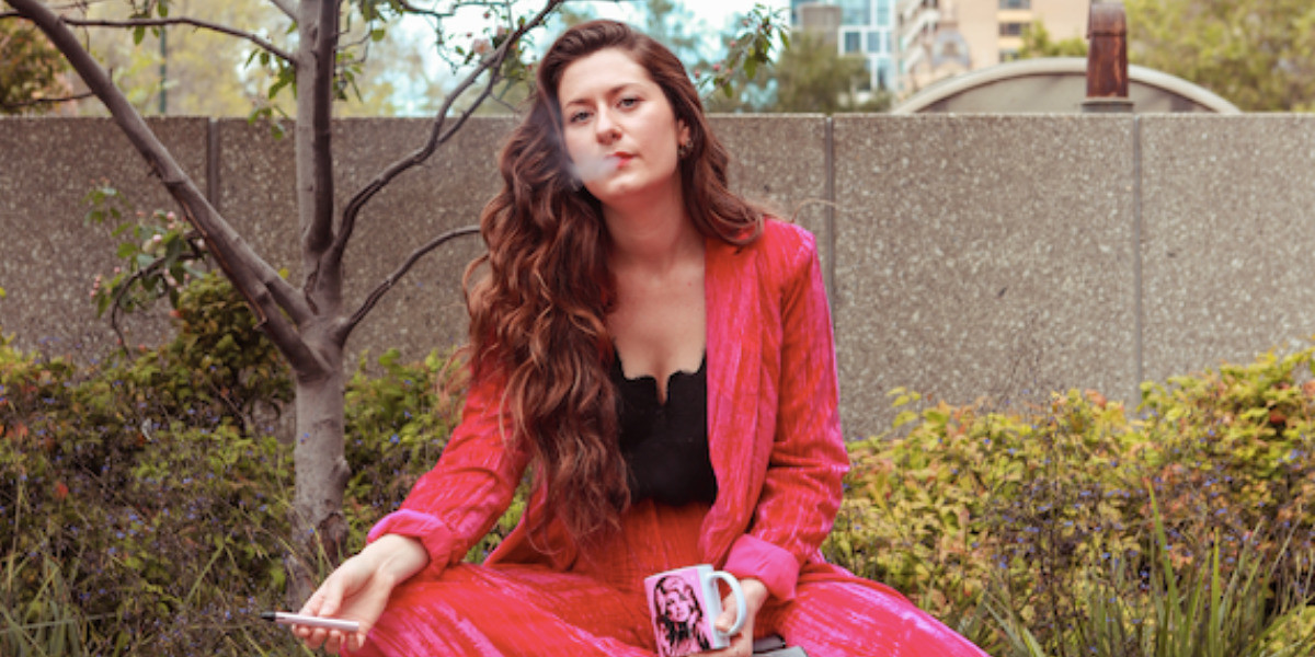 Image of young woman wearing a pink velvet suit, holding a pink Dolly Parton mug, and vaping.