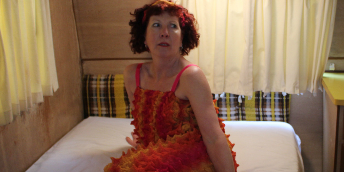 A woman dressed in a multi-coloured pointy-textured jumpsuit sits on a bed in a caravan. There are dark pink straps over her shoulders and she wears a pink and yellow headband in her short red curly hair. Her legs are folded in as she sits on her right hip supported by her right arm behind her. She is looking to the left with a scared expression on her face. The wall behind has a brown and yellow checked head rest below timber veneer panelling, and a cream curtain covers a window.