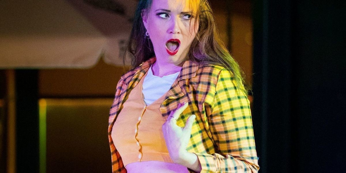 Brunette burlesque performer posed on stage wearing a yellow blazer and skirt that pays tribute to the costume worn by lead character in the movie 'Clueless'.