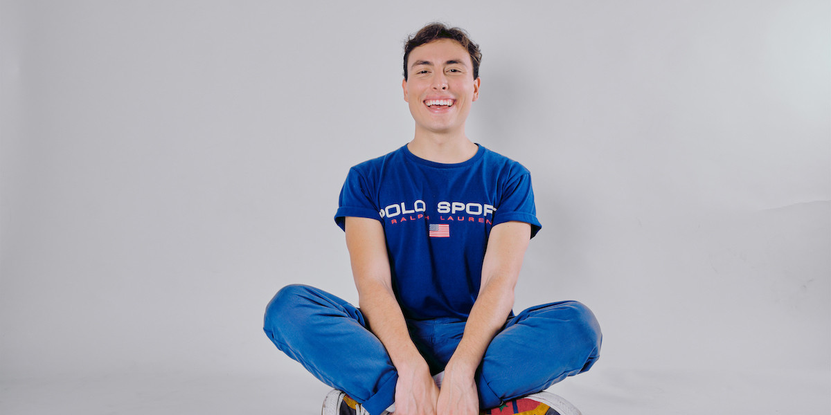 John is sitting cross-legged in front of a white background with a big smile on his face. He is wearing a matching bright blue t-shirt and blue pants.