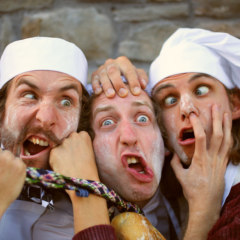 A close up photograph of three people with disgruntled facial expressions. Two people are wearing white baker’s hats, and each person has white baking flour on their faces. The person on the left has a fist punching their cheek and the person on the right has a finger up their nose.