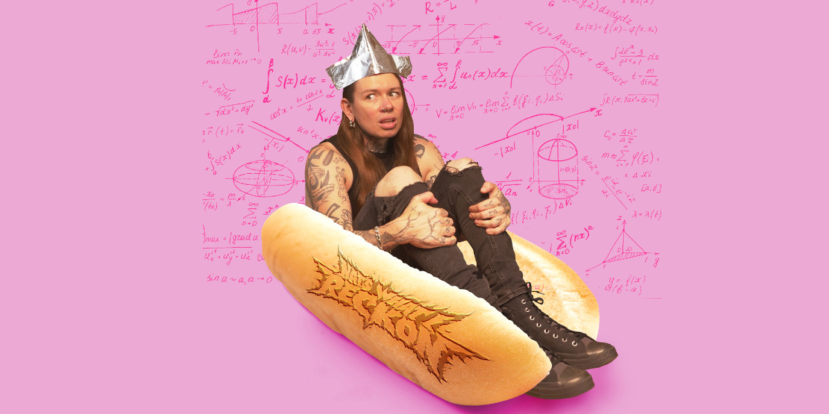 Nat has long hair, loads of tattoos & is wearing black jeans ripped at both knees, and a tinfoil crown. He is clutching his knees with both hands and looking off to his right with the expression of someone who doesn't trust what they are looking at. Maybe the expression is because he is wedged in a hot dog bun.