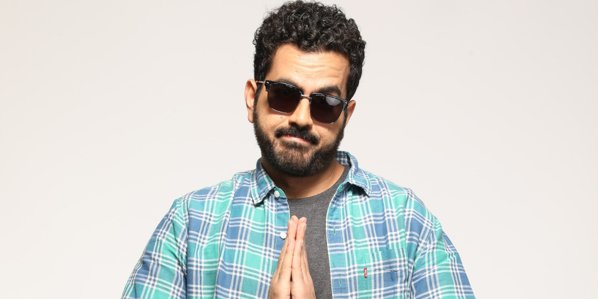 Performer wearing a blue chequered shirt, unbuttoned with a grey T shirt inside, wearing sunglasses, posing in the 'Namaste' sign