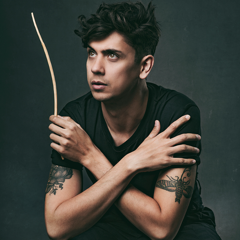 A performer with dark hair and green eyes, is sat cross legged with their arms crossed. In his left hand, he holds a wand pointing upwards.