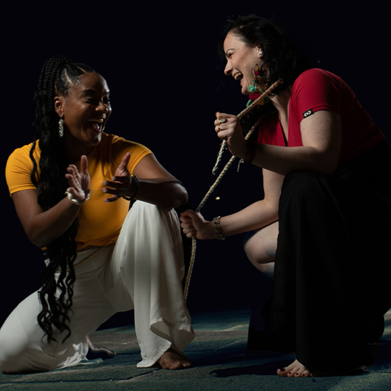 Two women laughing together while crouched on the ground.  The one on the left is clapping and the one on the right holds a rope that drapes around the back of her shoulders.