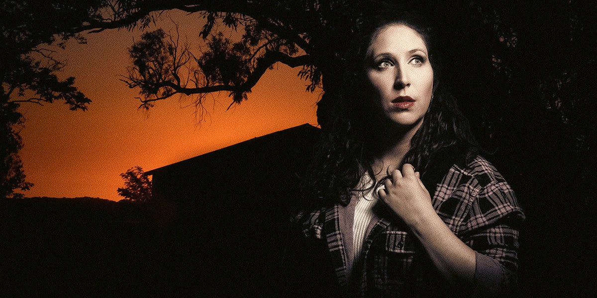 After Rebecca - A fair-skinned, brown-haired woman wearing a flannelette shirt stares out past the camera nervously. Behind her is the silhouette of a gum tree and a house against an orange sky.
