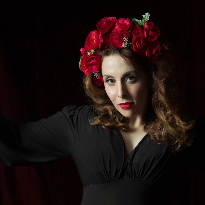 Euromash -  Johanna Allen. - A photograph of a woman seductively staring straight ahead. She is wearing a black long sleeved top and red lipstick. She has a headband made up of multiple red roses.