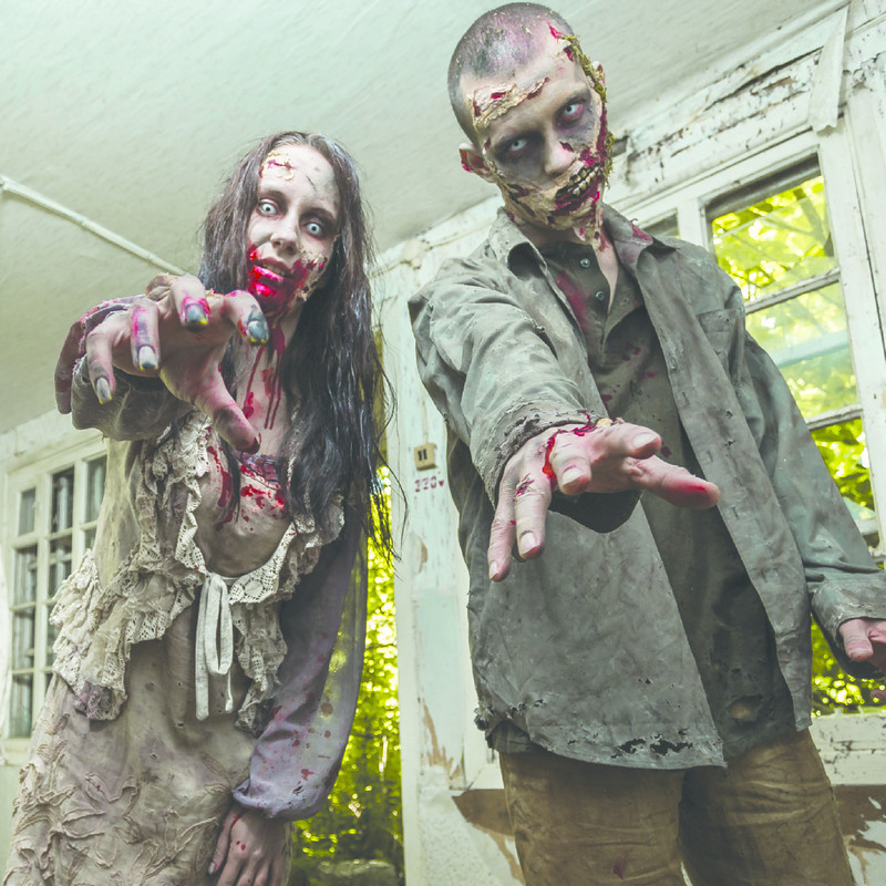 Two people dressed in tattered clothing, covered in blood with white eyes and peeling skin reach towards the camera. They are in a room with white peeling paint, there is green foliage through the windows and doorway behind them.