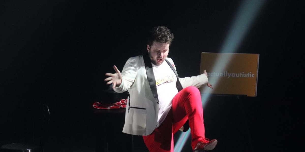 A white man with short dark curly hair is wearing a white shirt with Bazinga on the front, an off white Jacket with Black trim and red trousers. He has his head down, mouth open, and hands out to the side with fingers spread. He has one knee bent and raised in what looks like a high marching pose.