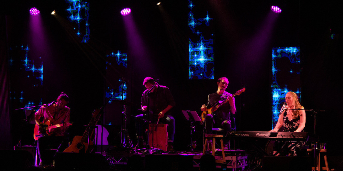 Four musicians are playing on a stage. Left to right they are guitarist, percussionist, bass guitar and keys/singer. There is blue patterned stage lighting behind them and three purple spotlights shining on them from above.