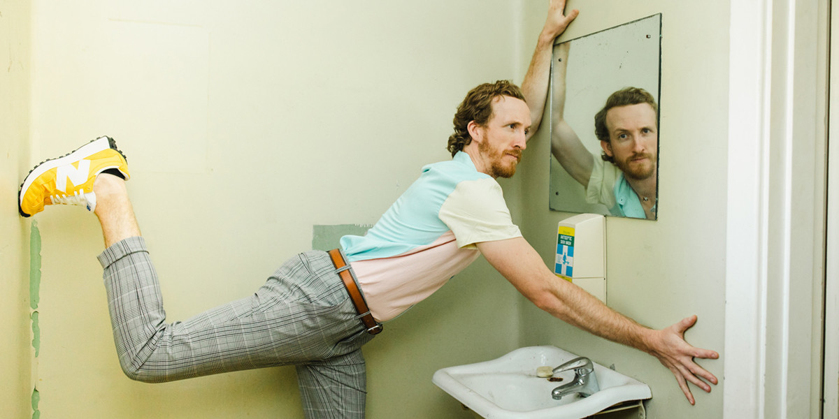 Will is squished into an awkward position in a grungy bathroom. His hands are over a dirty sink, and he's looking at the camera through the reflection of a mirror. He has a quizzical look on his face, and is wearing a pastel color shirt, bright yellow shoes, and striped grey suit pants.