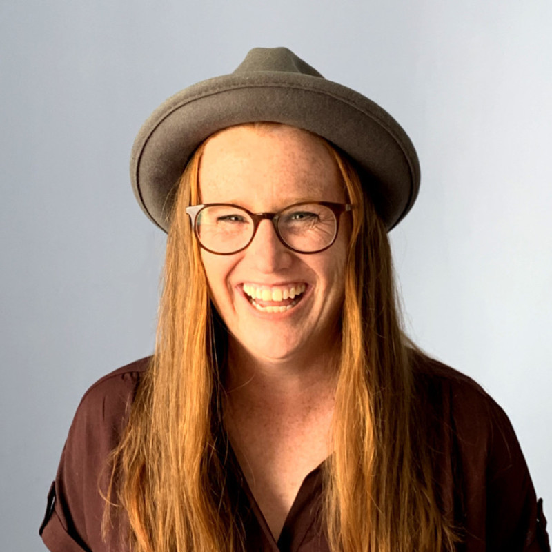 A photo of a person with long blonde hair laughing. They are wearing a dark grey felt hat and a brown shirt.