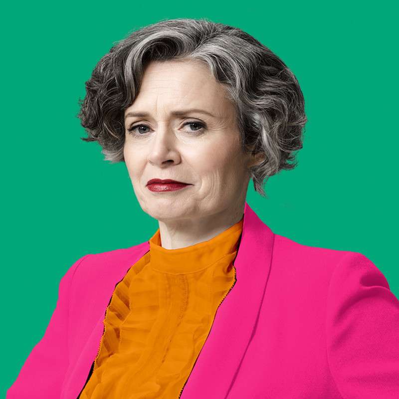 JUDITH LUCY - TURNS OUT, I'M FINE: IN CONVERSATION - Judith has short wavy silver and black hair. She is wearing a bright pink suit jacket and bright orange frilly blouse. She is wearing dark red lipstick and has a slight smirk on her face. She is standing in front of a dark green background.