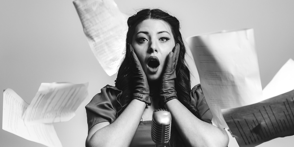 Great Detectives: All New Mysteries! - Black and white photo. A woman (Tate Simpson), dressed in 1950s attire, gasps in shock while standing at a vintage microphone while script pages cascade around her.