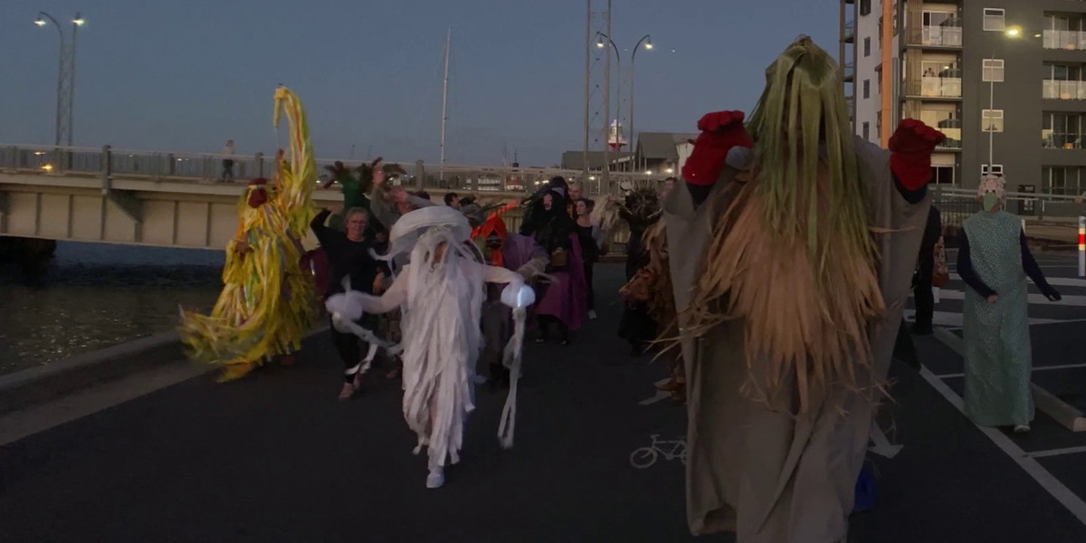 A procession of strange masked figures is walking along the dock by the Port River. In the foreground one of the figures is a 10 foot green grey cloaked figure with a stylised palm frond as a face and gigantic red hands raised like a Bear.