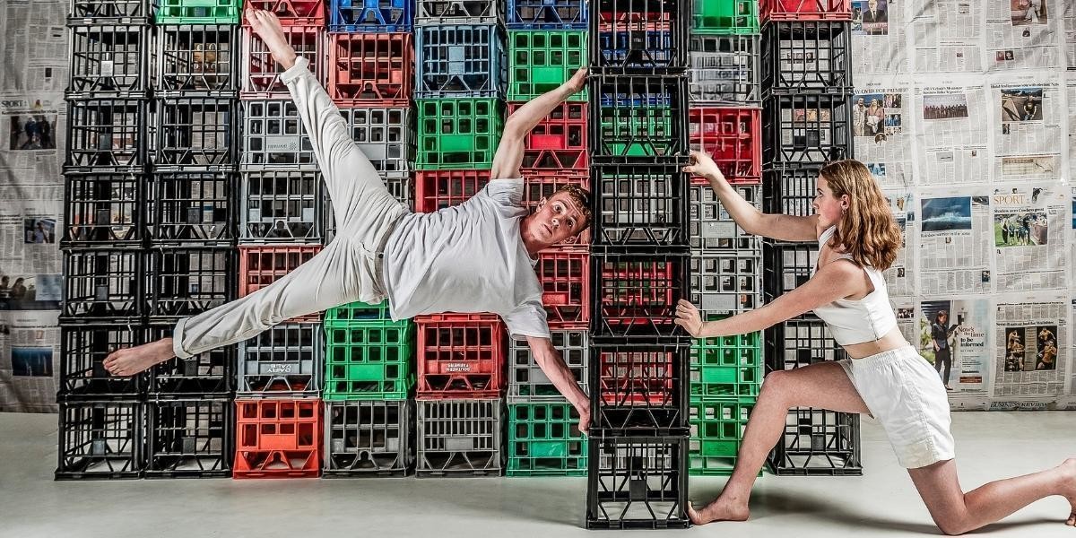 A teenage male acrobat dressed in white holds a 'flag' position while holding onto a tall stack of milk crates. A teenage female acrobat holds the stack from the other side. They are in front of a wall of milk crates.
