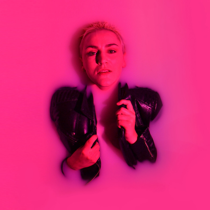 Grief Lightning: A Satire In 78 Slides - A figure is on their back partly submerged in pink milky liquid, their hands, face and parts of their leather jacket are exposed above the liquid. The figure has short blond hair and fair skin, a pink light is covering the entire image.