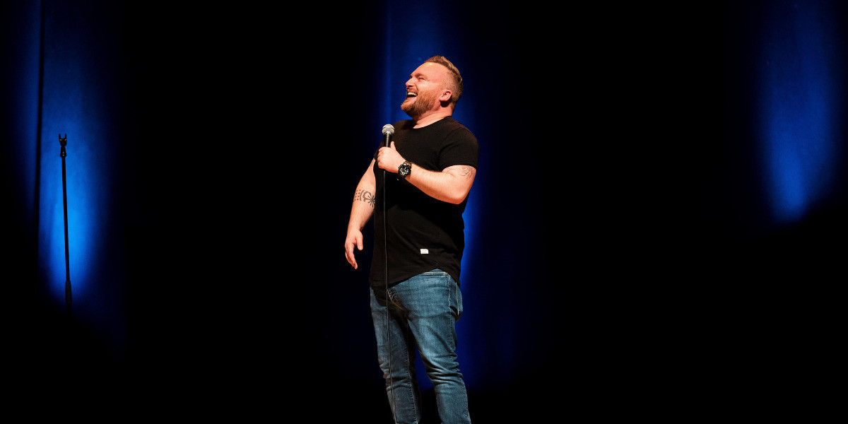 Micky Bartlett - THICC - Man stands onstage with a microphone in hand throwing head back in laughter