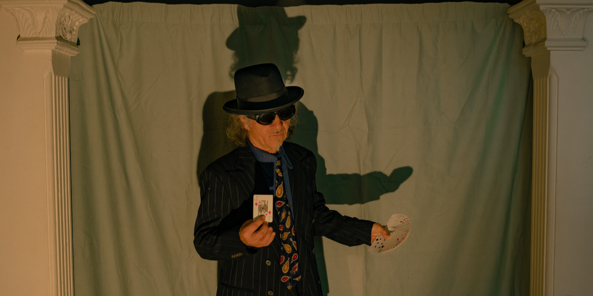 A man wearing a suit, sunglasses and a hat, holding a fan of playing cards in one hand and the jack of diamonds in the other.