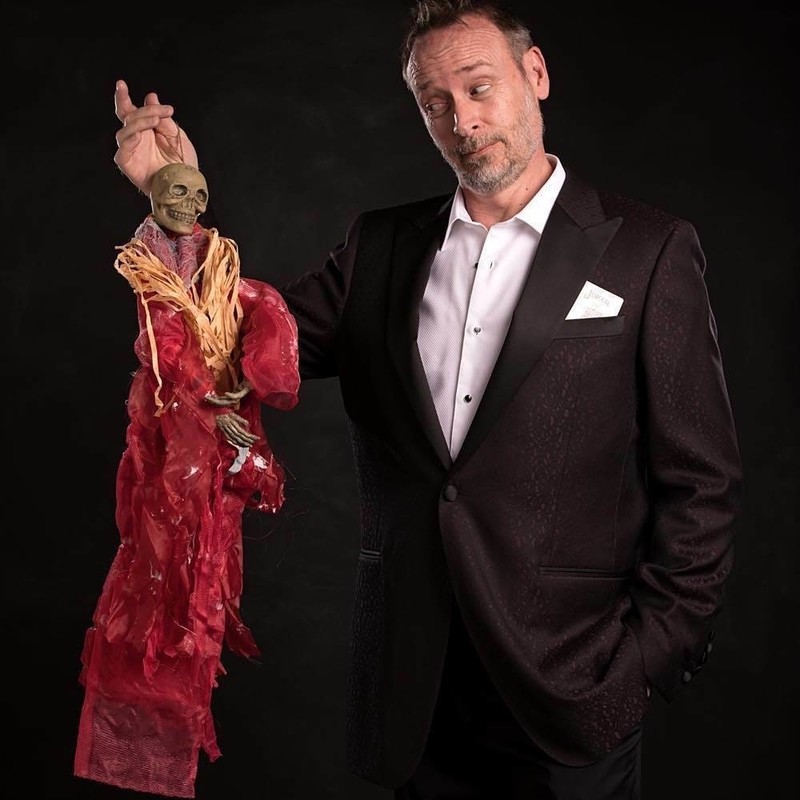 A man holds a skeleton puppet that wears a long red cloak. The man wears a black dinner suit and white shirt, he has a card poking out his jacket pocket. He is looking suspiciously at the puppet.