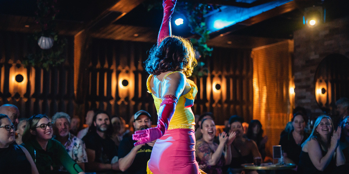 Brunette burlesque  performing a sixties Gogo dance move on stage, wearing a yellow and pink dress and top.