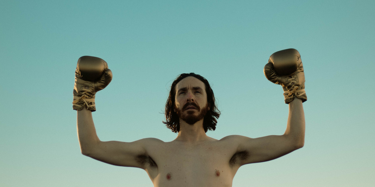 A bearded man stands shirtless with golden boxing gloves on in front of a pale blue sky