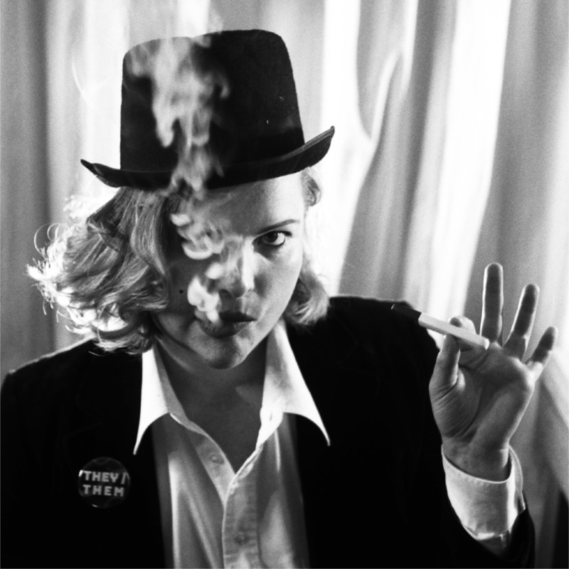 Them Fatale - Black and white photo of James Penwarden wearing a suit, "they/them" badge, and black hat. They exhale vape haze in the style of Marlene Dietrich.