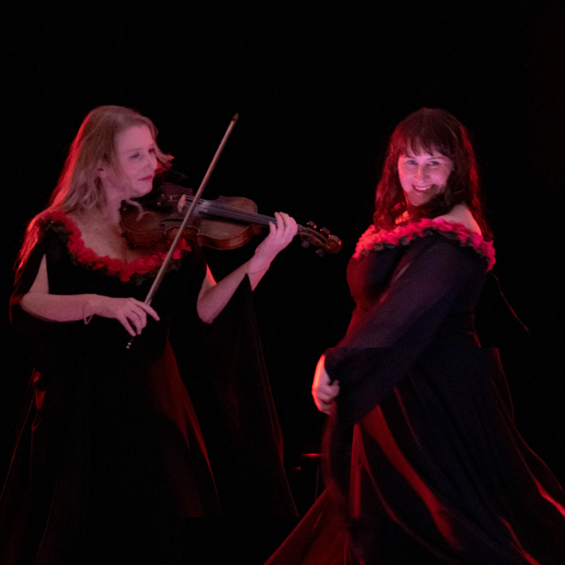 A blonde woman plays the violin while a brunette looks at the camera whilst singing and dancing
