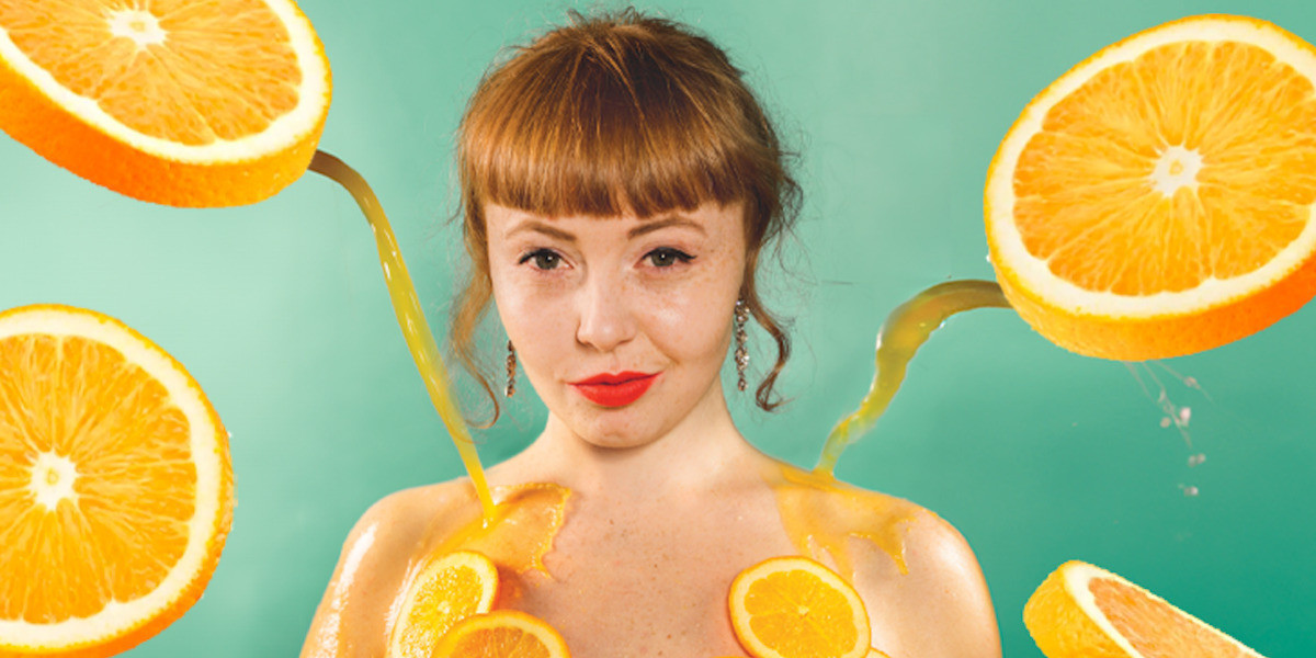 Fruition - A woman with red hair and orange-red lipstick stares at the camera in a sultry way, while orange juice pours towards her.