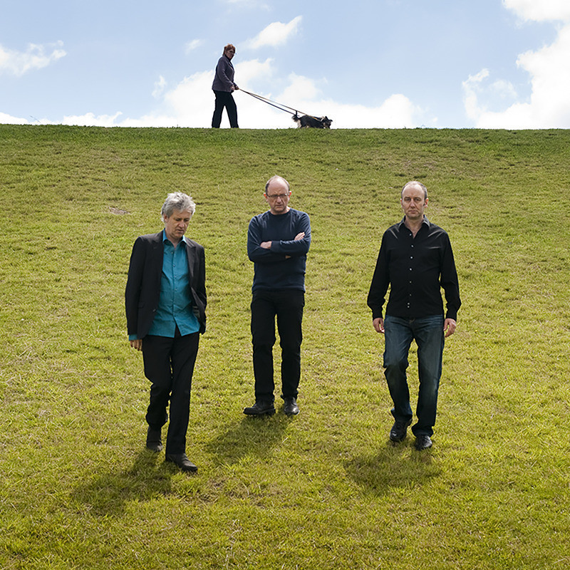 Three men in dark clothing walking down a grassy hill towards camera on a sunny day with blue sky and clouds. A person pushing a lawn mower at top centre of hill behind them.