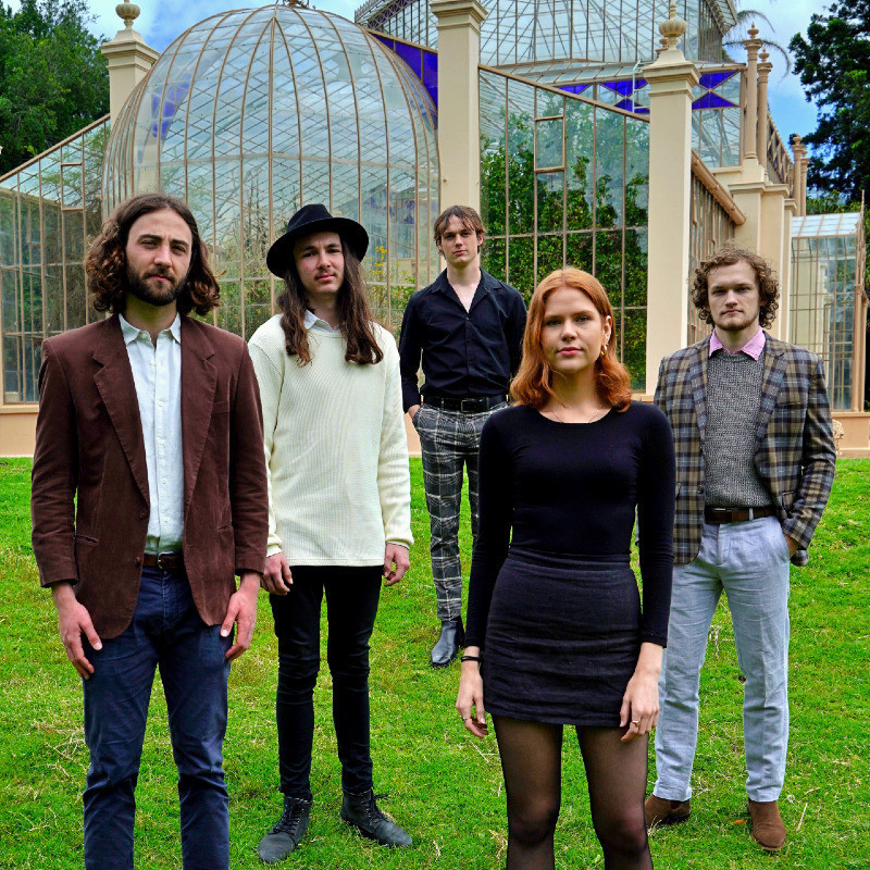 Five people posed on a grassy hill in front of a historic greenhouse covered in glass panes. The person at the front has long red hair and is wearing a black shirt with a grey skirt and black leggings. Everyone in the photo has a serious, inquisitive look on their face.