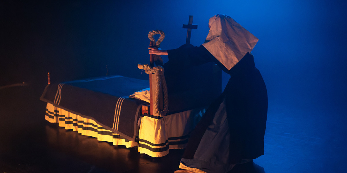 Woman dressed as nun has bakc to us and pulls a bed (or is it a boat?) across the stage