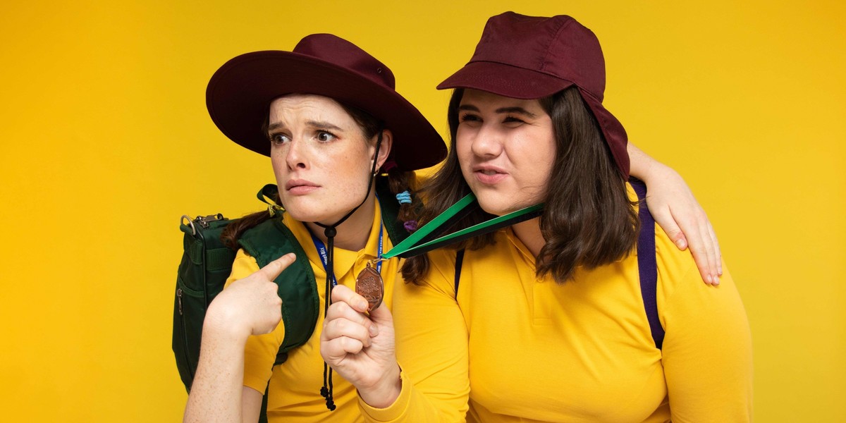 Two white women in yellow school polo shirts and burgundy school hats stand with arm around each other. They are holding a participation medal with a 'so there!' look on their faces.