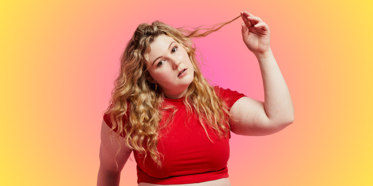 Tori, a white woman with curly blonde hair, lifting a piece of her hair loftily in her  hand and looking at the camera.