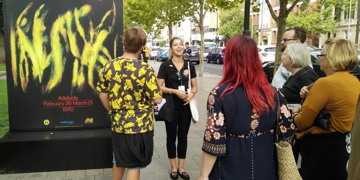 Adelaide Fringe: A History - A tour guide talks to guests standing next to a display poster from the Adelaide Fringe.