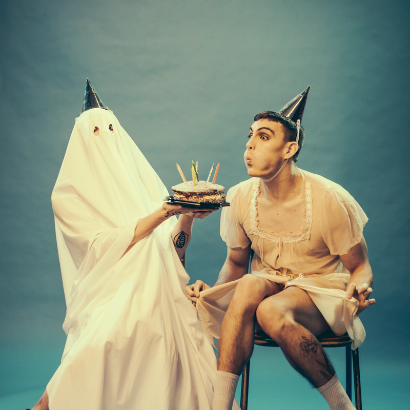 Two performers are seated slighting facing in toward each-other. One performer is dressed in a ghost sheet wearing a party hat, red high heels and holding a cake, the other is wearing a tan nighty, party hat and black high heels. The performer in the tan nighty is pretending to blow out a candle atop the cake.
