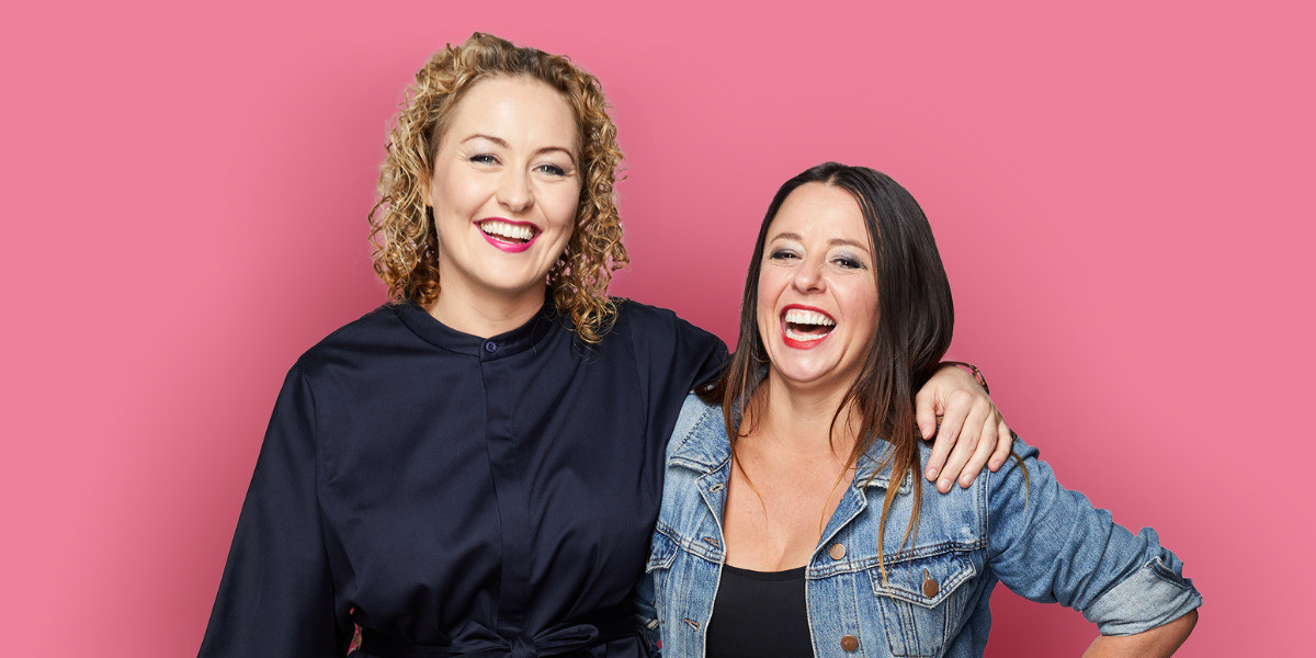 Bang On Live - Myf Warhurst & Zan Rowe - From left to right: Zan has short blond curly hair. She is wearing a navy-blue dress. Myf has long brown straight hair, she is wearing a black top and blue denim jacket. They are both laughing and looking at the camera. The background is pink.