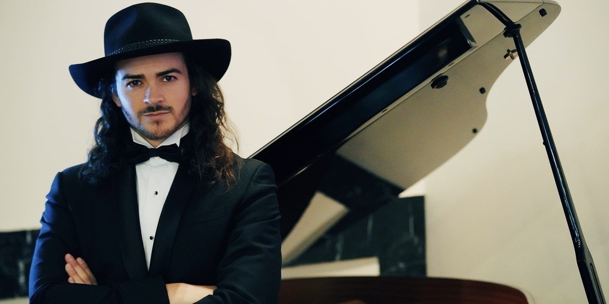 Chooka Parker looking dapper in a Tuxedo & black Akubra hat leaning against a grand piano.