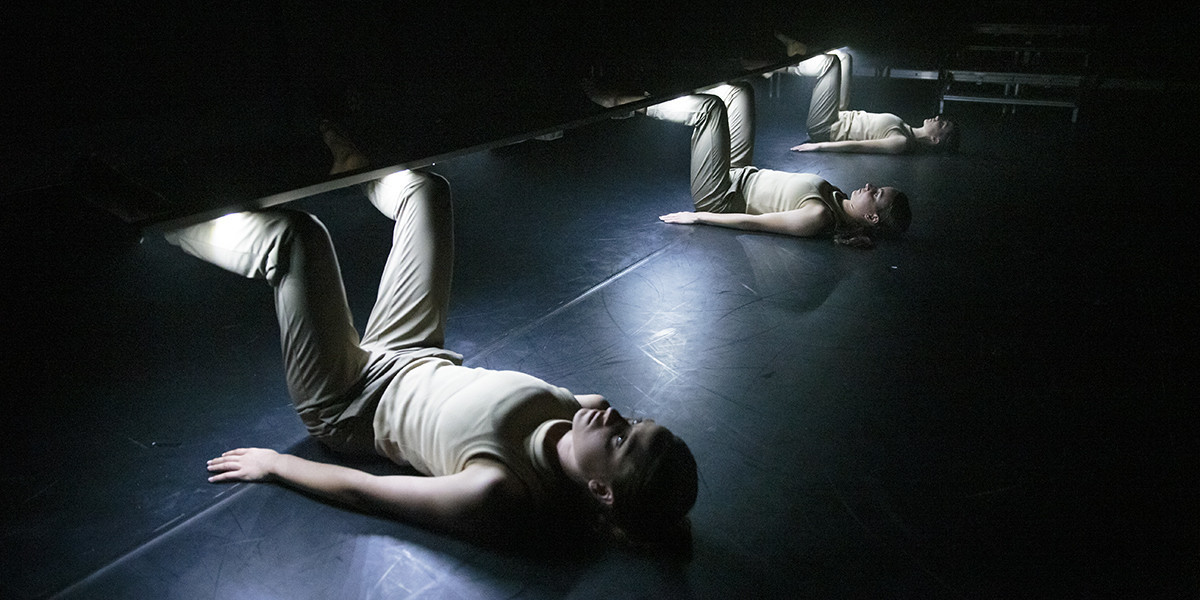 A row of three performers lying on a dark stage with their knees raised in the air. Resting on their shins is a long plank from which bright white lights shine down onto their bodies.