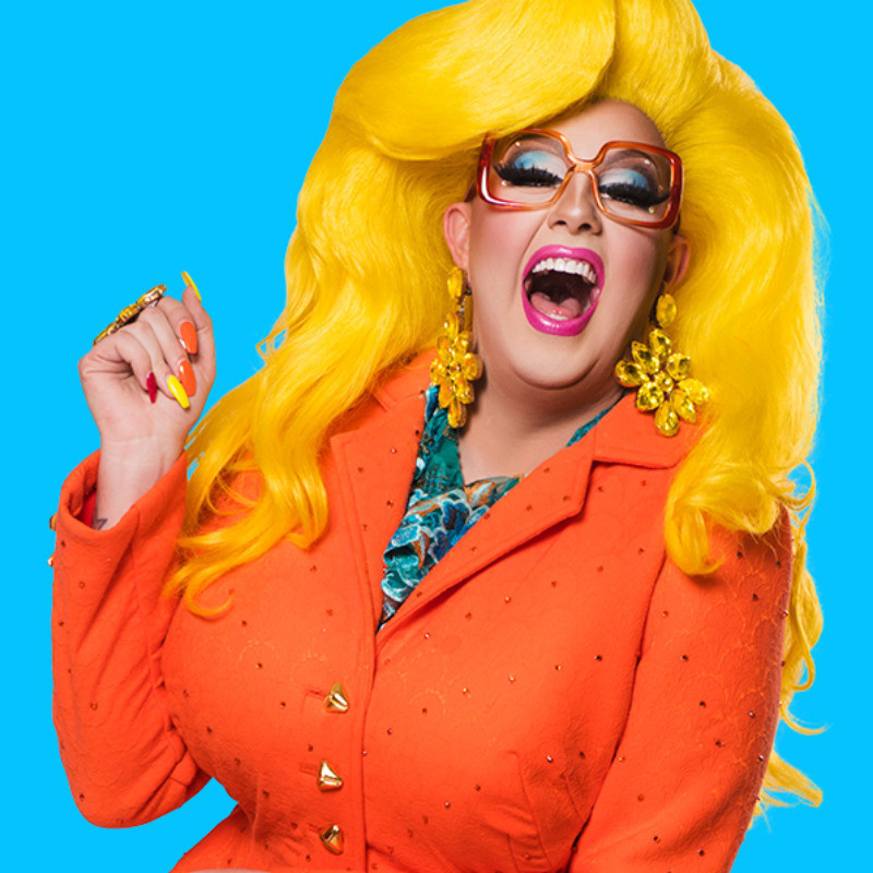 Karen From Finance is Out Of Office - A photo of a person laughing. They have voluminous bright yellow hair and they are wearing a bright orange jacket. They are wearing big yellow earrings and orange glasses.