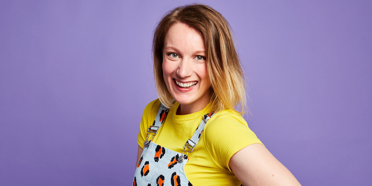 Eleanor is smiling at the camera wearing a yellow t-shirt and a denim pattered overalls standing in front of a purple backdrop.